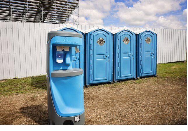 Portable restrooms placed along a public park path near the water, with the city skyline visible in the background